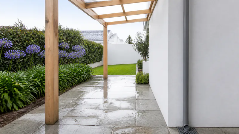 A patio of wet grey flagstones under a timber pergola with a clear roof, beside a white house wall where a grey downpipe runs down into a square grated drain set in the paving
