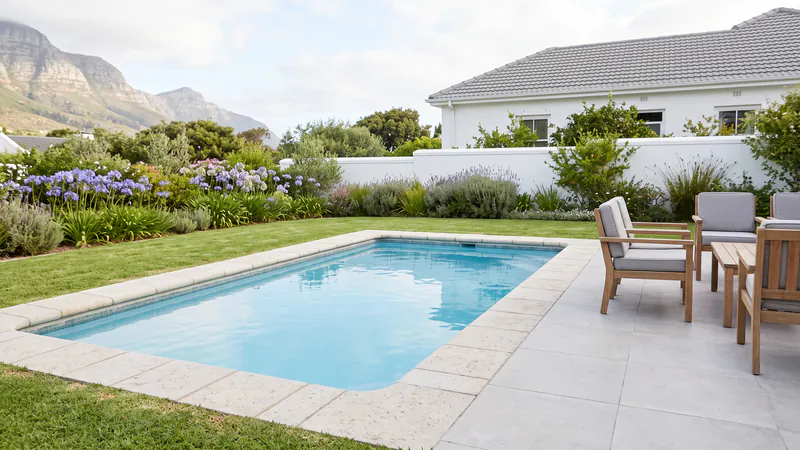 A rectangular swimming pool edged with pale stone coping, continuing into a patio of large pale-grey tiles with wooden outdoor chairs, lawn and agapanthus around it