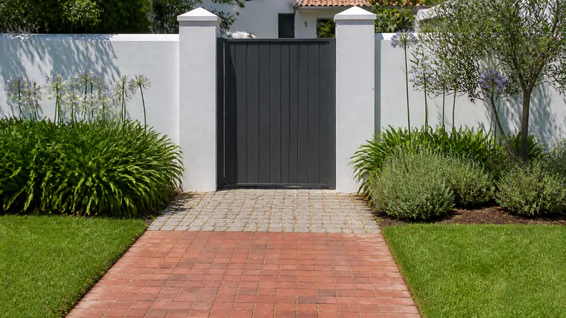 A garden path of red-brown clay pavers between two lawns, meeting a small apron of grey granite setts in front of a dark garden gate set in a white wall