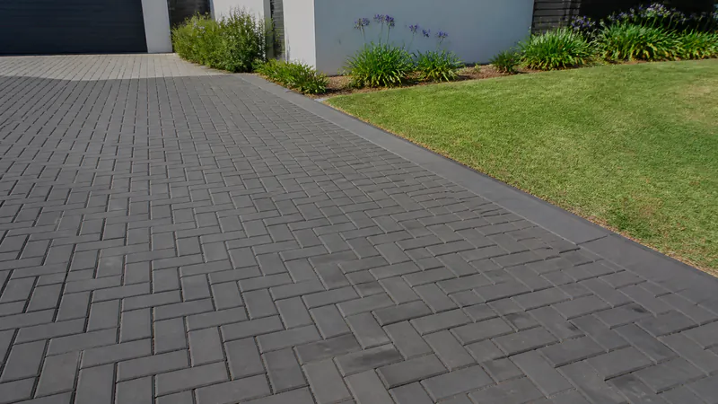 A driveway of plain rectangular charcoal concrete paving blocks laid in herringbone, with a straight dark edge strip along a lawn and agapanthus in front of a white wall