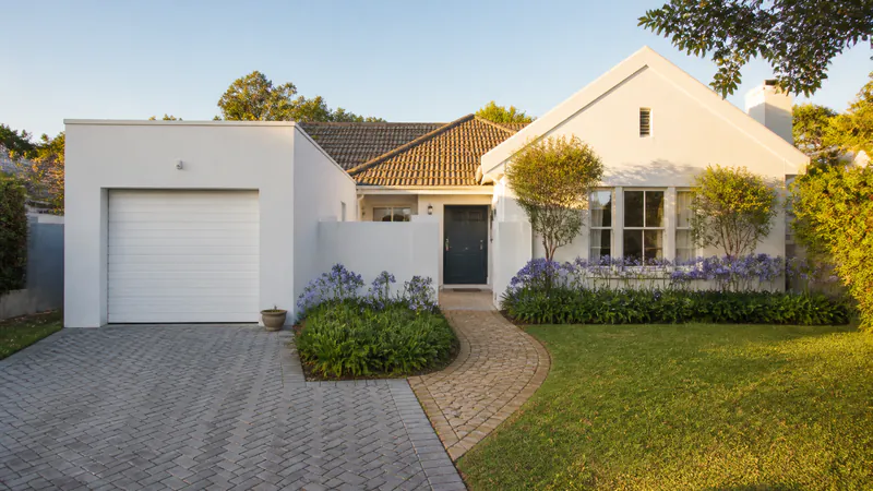 A white house with a grey herringbone block-paved driveway to a white garage door, and a separate curving path of tan cobbles across the lawn to the dark front door